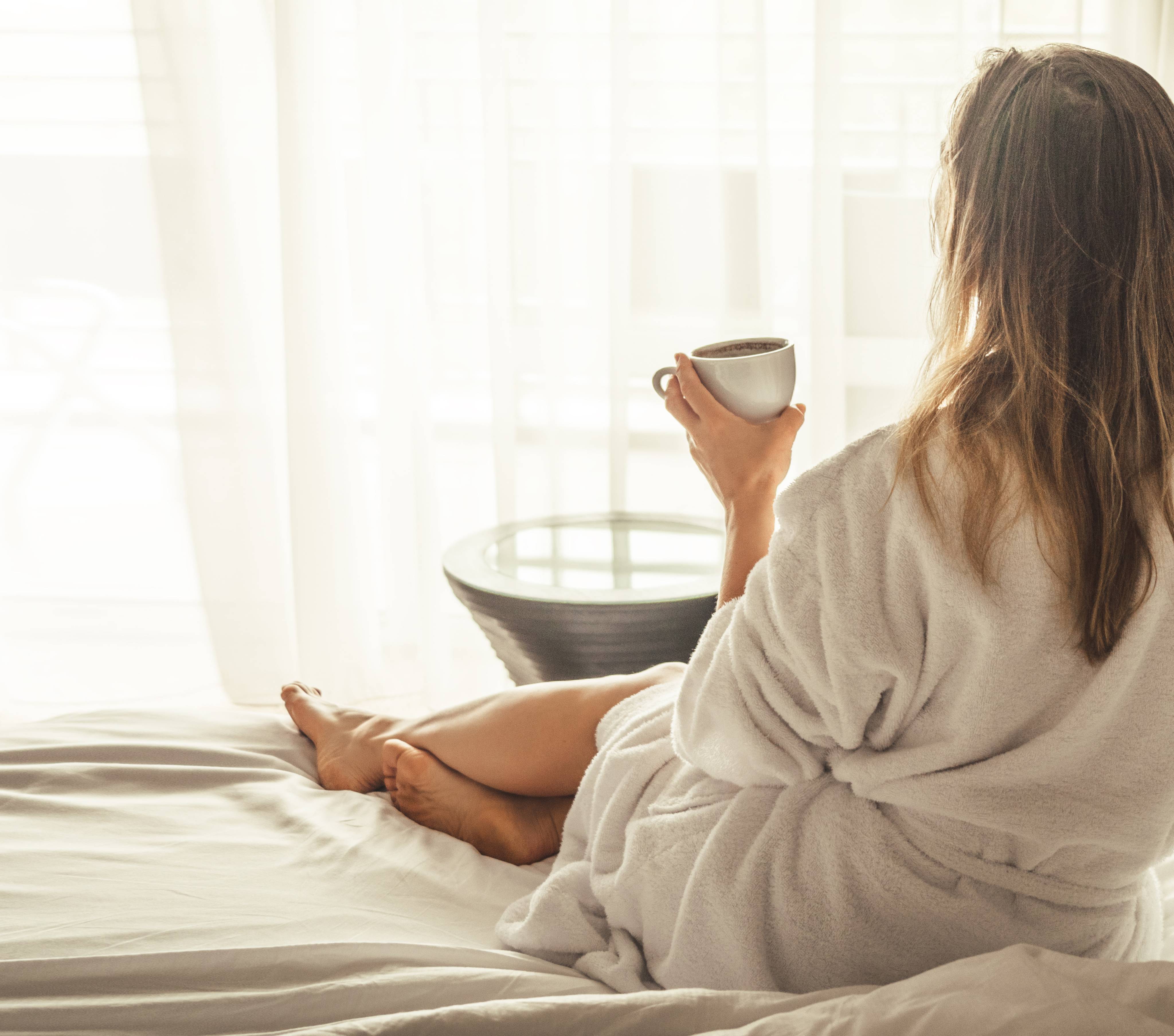 Frau im Bademantel sitzt auf dem Bett im Hotelzimmer und trinkt einen Kaffe